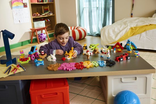 Een jongen met een paarse trui bouwt met LEGO-stenen op een tafel, omringd door diverse creaties.