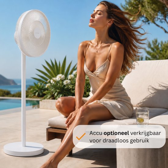 Een vrouw met gesloten ogen zit naast een witte staande ventilator op een loungebank bij een zwembad. "Accu optioneel verkrijgbaar voor draadloos gebruik".