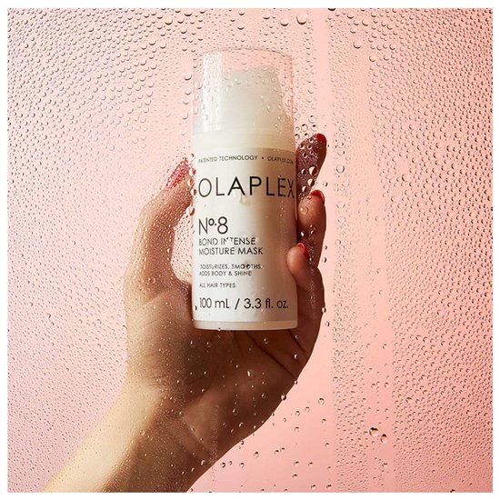 Hand houdt witte OLAPLEX Nr. 8 fles vast tegen een roze achtergrond met waterdruppels.