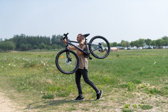 Man met een zwarte mountainbike over zijn schouder, lopend door een veld.