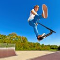 Jongen met rode haren maakt een sprong in de lucht met een zwarte stuntstep, op een zonnige dag in een skatepark.