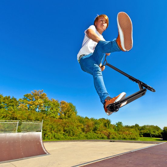 Jongen met rode haren maakt een sprong in de lucht met een zwarte stuntstep, op een zonnige dag in een skatepark.