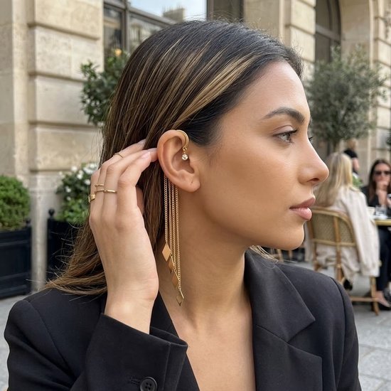 Model draagt een goudkleurige ear cuff met pareldetail en lange kettingen met ruitvormige hangers. Ze draagt een zwarte blazer.