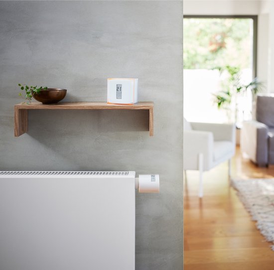 Netatmo slimme thermostaatknop op witte radiator. In de achtergrond een plank met thermostaat en zithoek.