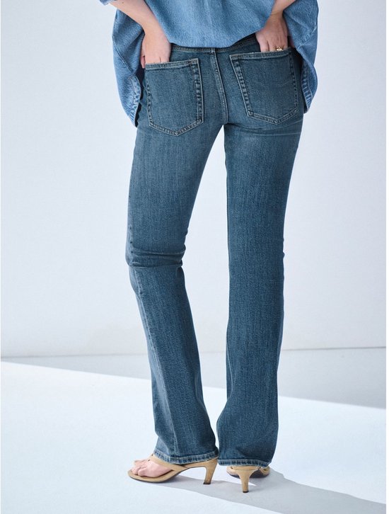 Zijaanzicht van model dat een spijkerbroek met bootcut pijpen draagt, met de handen in de zakken.