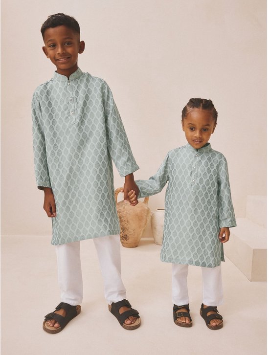 Twee jongens die elkaars hand vasthouden, gekleed in lichtgroene kurta's met bladpatroon en witte broeken.