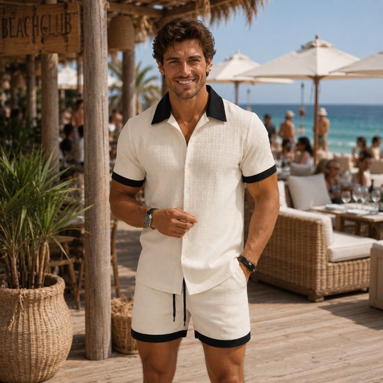 Man in off-white linnenlook set, lachend op een strandlocatie. Zwart kraagje en boorden.