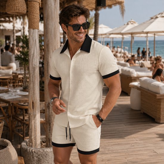Man in off-white linnenlook set, lachend op een strandlocatie. Zwart kraagje en boorden.