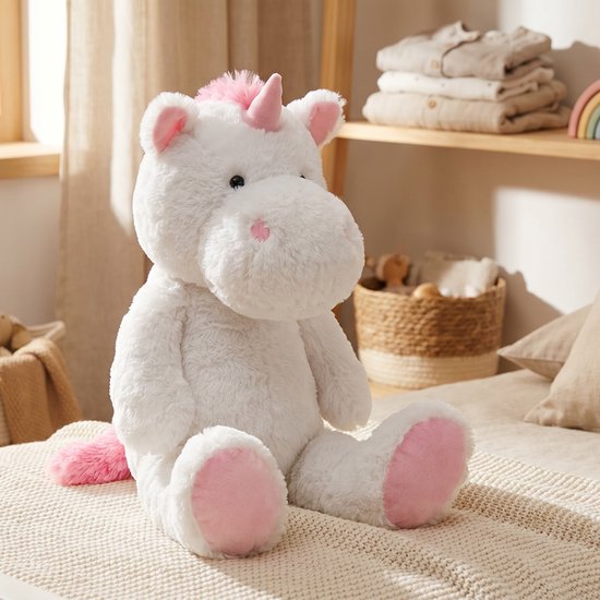 Pluche eenhoorn knuffel, wit met roze details zoals hoorn, oren, staart en voetzolen. Zit op een bed.
