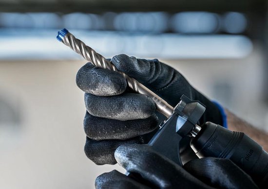 Een persoon met zwarte handschoenen houdt een Bosch SDS plus-7X hamerboor vast in een boorhouder.