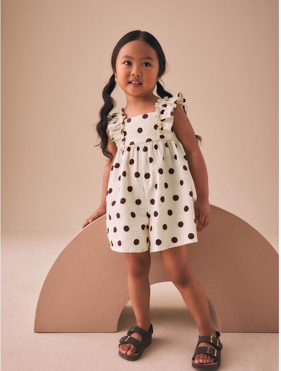 Meisje met staartjes draagt een crèmekleurige playsuit met bruine stippen, ruches en sandalen.