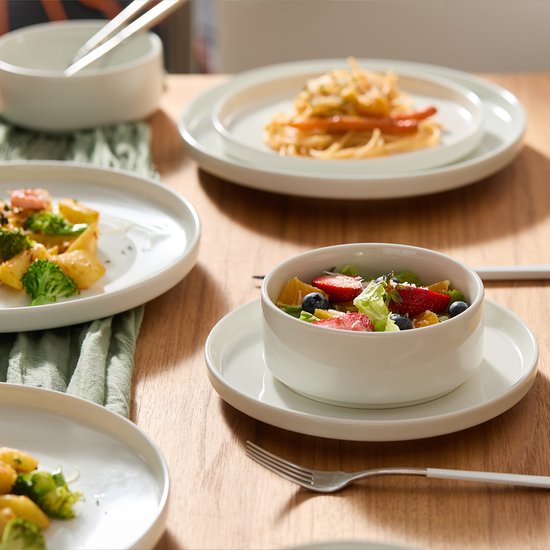 Een gedekte tafel met witte borden en kommen, een kom met fruitsalade, borden met pasta en salade.