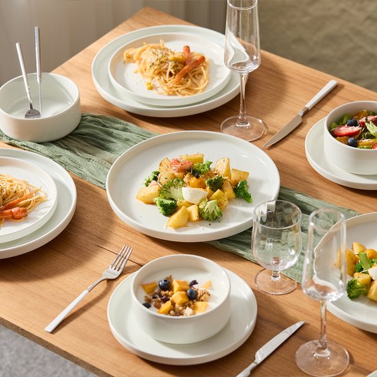 Een gedekte tafel met witte borden en kommen, geserveerd met pasta, salade, en yoghurt met fruit.