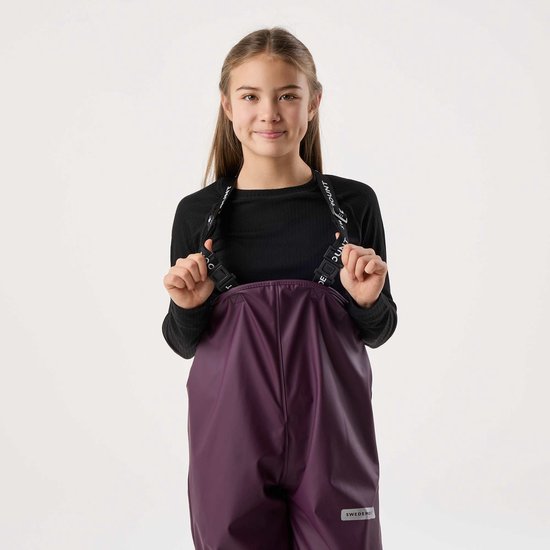 Meisje met lang bruin haar, draagt een zwarte top en paarse bretelbroek. Logo op de banden.