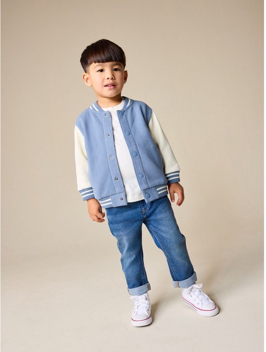 Kind in een blauw-wit bomberjack, wit shirt, jeans en witte sneakers.