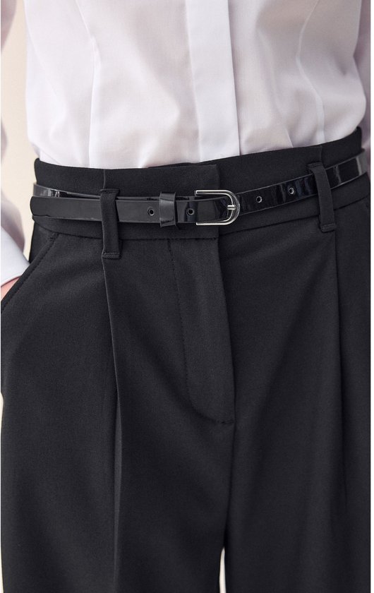 Meisje draagt een zwarte wijde broek met riem en wit overhemd, handen in zakken.