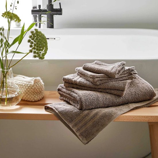 Stapel taupe handdoeken met ribbelrand, bij een bad en vaas met groen blad.