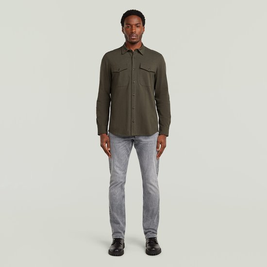 Man met donkergroen overhemd met lange mouwen, twee borstzakken en grijze jeans.