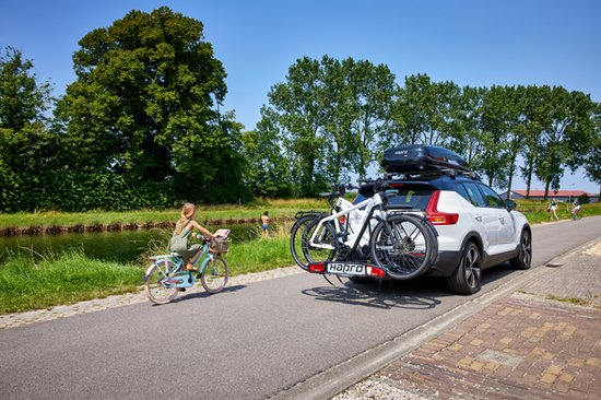 Witte auto met dakdragers en een fietsendrager met twee fietsen erop, geparkeerd bij een water met een fietser op de voorgrond.