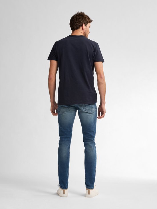 Man met kort bruin haar, staand van achteren gezien, draagt een marineblauw t-shirt en een vaalblauwe slim fit jeans.
