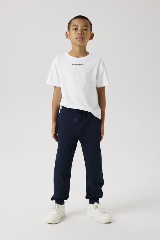 Jongen in een witte T-shirt met tekst 'Studio Edition' en een donkerblauwe joggingbroek, met witte sneakers.
