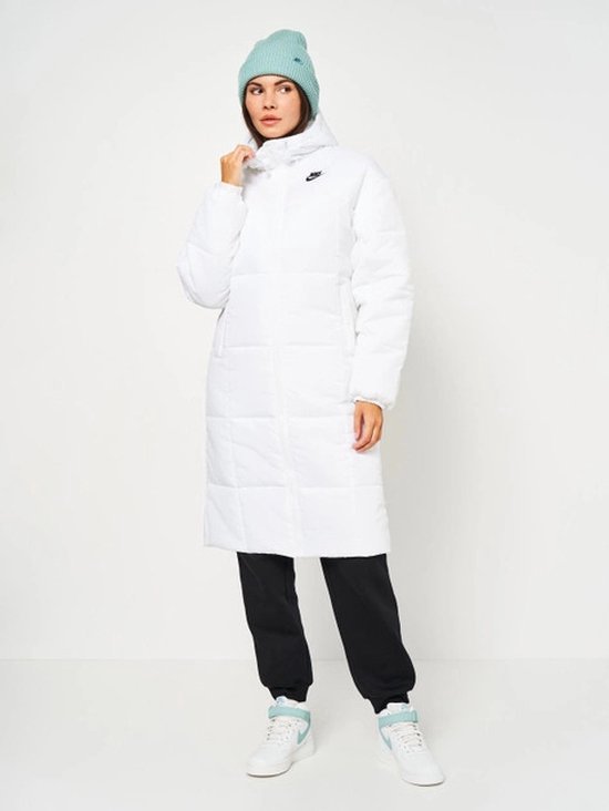 Wit, lang Nike winterjack met capuchon, gedragen door een model met een lichtblauwe muts en zwarte broek.