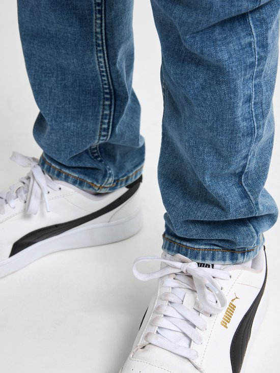 Blauwe jeans met witte Puma sneakers. De broekspijpen vallen over de schoenen.