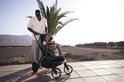 Een man duwt een donkergroene kinderwagen met een kindje erin op een betegeld terras.