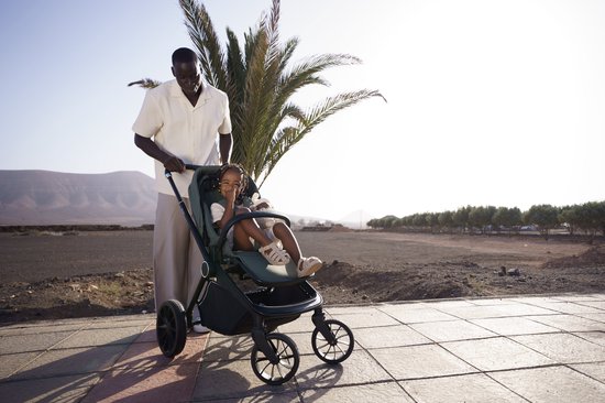 Een man duwt een donkergroene kinderwagen met een kindje erin op een betegeld terras.