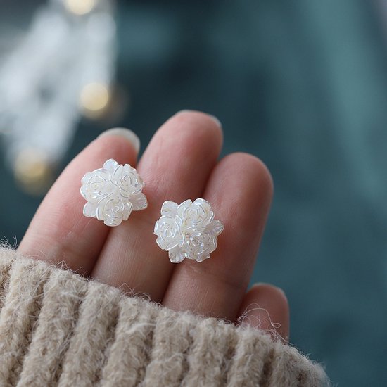 Twee witte bloem-vormige oorknoppen met gedetailleerde blaadjes, vastgehouden in een hand met een gebreide trui.