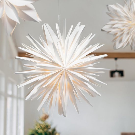 Witte papieren ster met veel punten en warm licht, hangend in een lichte kamer met kerstboom.