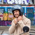 Man met donkerblauwe helm met vizier en beige jas, die de kinband vastklikt.