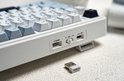 Detail van de zijkant van een wit mechanisch toetsenbord met USB-C poort, aan/uit-schakelaar en USB-dongle.