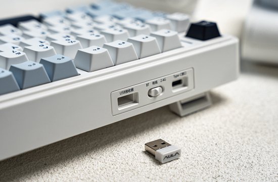 Detail van de zijkant van een wit mechanisch toetsenbord met USB-C poort, aan/uit-schakelaar en USB-dongle.