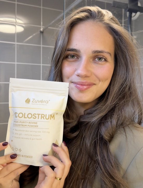 Jonge vrouw met lang bruin haar houdt een beige zak met "Zuvéra Colostrum" omhoog. Ze glimlacht naar de camera.