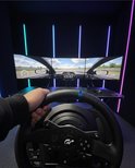 Een persoon houdt een race stuur vast in een opstelling met drie monitoren waarop een racegame wordt gespeeld.