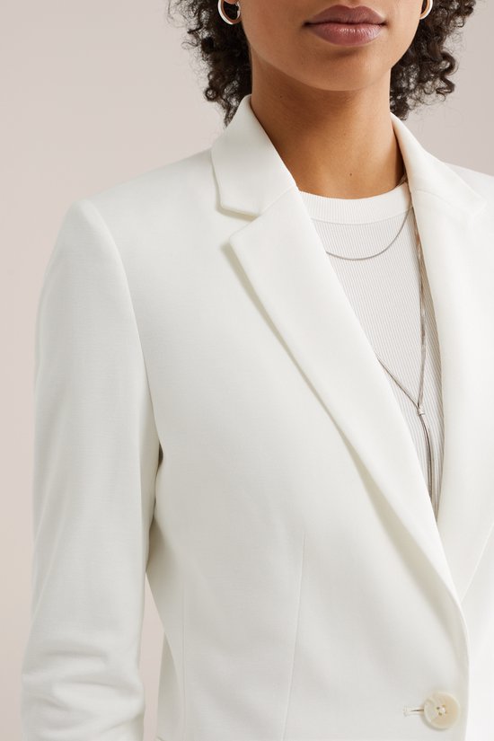 Close-up van een witte getailleerde blazer over een witte top met een zilverkleurige ketting.