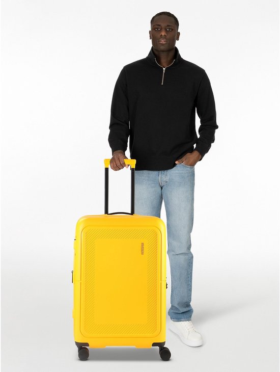 Man met zwarte trui en spijkerbroek staat naast gele American Tourister koffer.