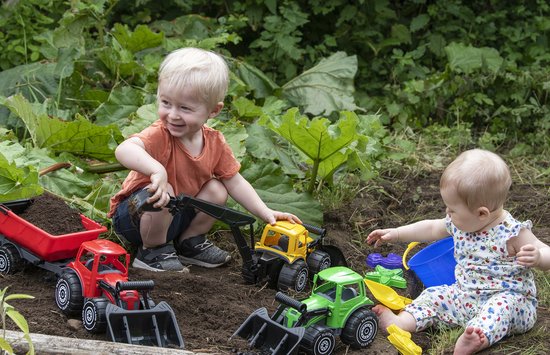 Blonde jongen en baby spelen buiten met speelgoed vrachtwagens: een rode kipper, gele graafmachine en groene tractor met aanhanger.