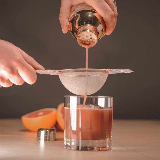 Cocktail wordt gefilterd door een zilverkleurige theezeef met decoratieve vleugels in een glas met ijs. Helft van een grapefruit op de achtergrond.