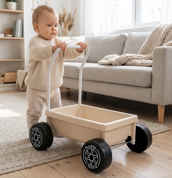 Een baby in een gebreide trui duwt een zandkleurige Plasto kinderwagen in een woonkamer.
