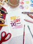 Close-up van een aquareltekening met de tekst "YOU CAN DO THIS", omringd door STABILO woody potloden.