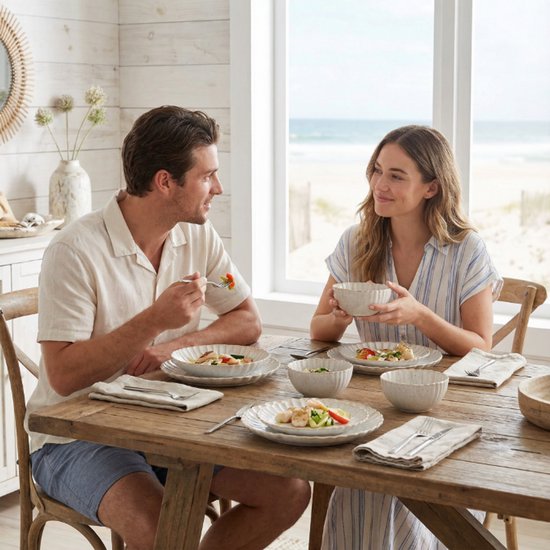 Een stel geniet van een maaltijd bij een houten tafel, zittend aan gedekte borden met gegolfde randen. Een strandtafelsetting.