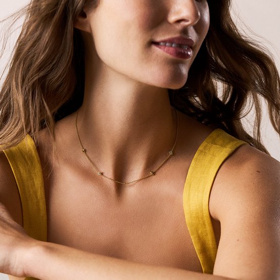 Gouden collier met vier kleine bedeltjes: twee hartjes en twee steentjes, gedragen door een vrouw met een gele top.
