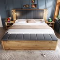 Houten bed met grijs gestoffeerd hoofdbord, 2 nachtkastjes met lampjes en opbergbank, bed is gedrapeerd met lakens.