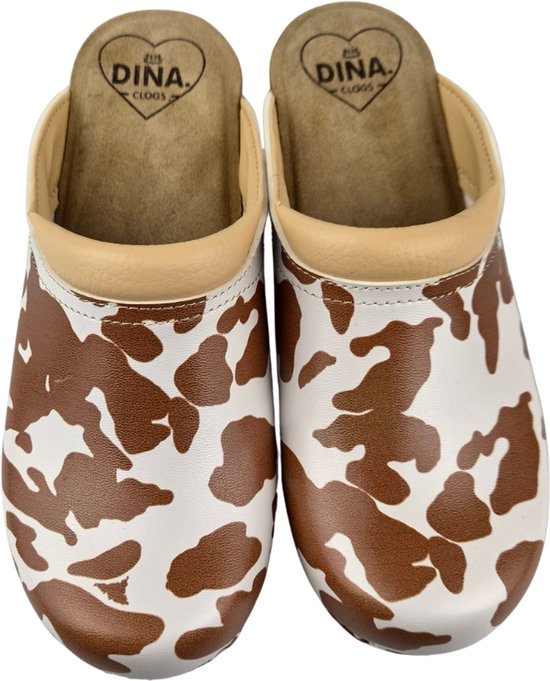 Een paar klompen van wit leer met bruine koeienvlekkenprint. Beige leren rand en binnenzool met 'DINA CLOGS' logo.