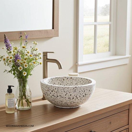 Terrazzo waskom met zwart-wit mozaïekpatroon op een houten badmeubel, naast een bos bloemen en zeepdispenser.