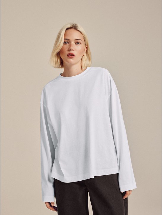 Model met blond haar, draagt een witte longsleeve met ronde hals en donkere broek.