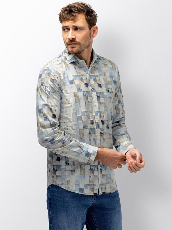 Man draagt een langarmig overhemd met een mozaïekprint van beige, blauwe en witte vierkanten, gecombineerd met spijkerbroek.