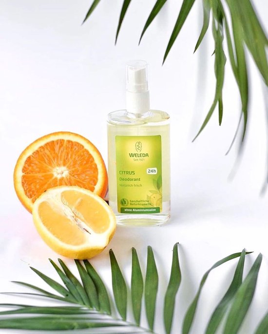 Weleda Citrus Deodorant Spray met een halve sinaasappel en een halve citroen op een witte achtergrond, omringd door groene bladeren.